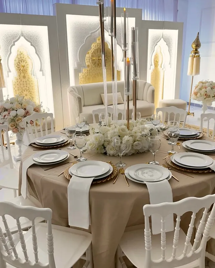 Table ronde nappée ivoire doré, assiettes blanches, couverts dorés et chandeliers de verre devant le canapé des mariés